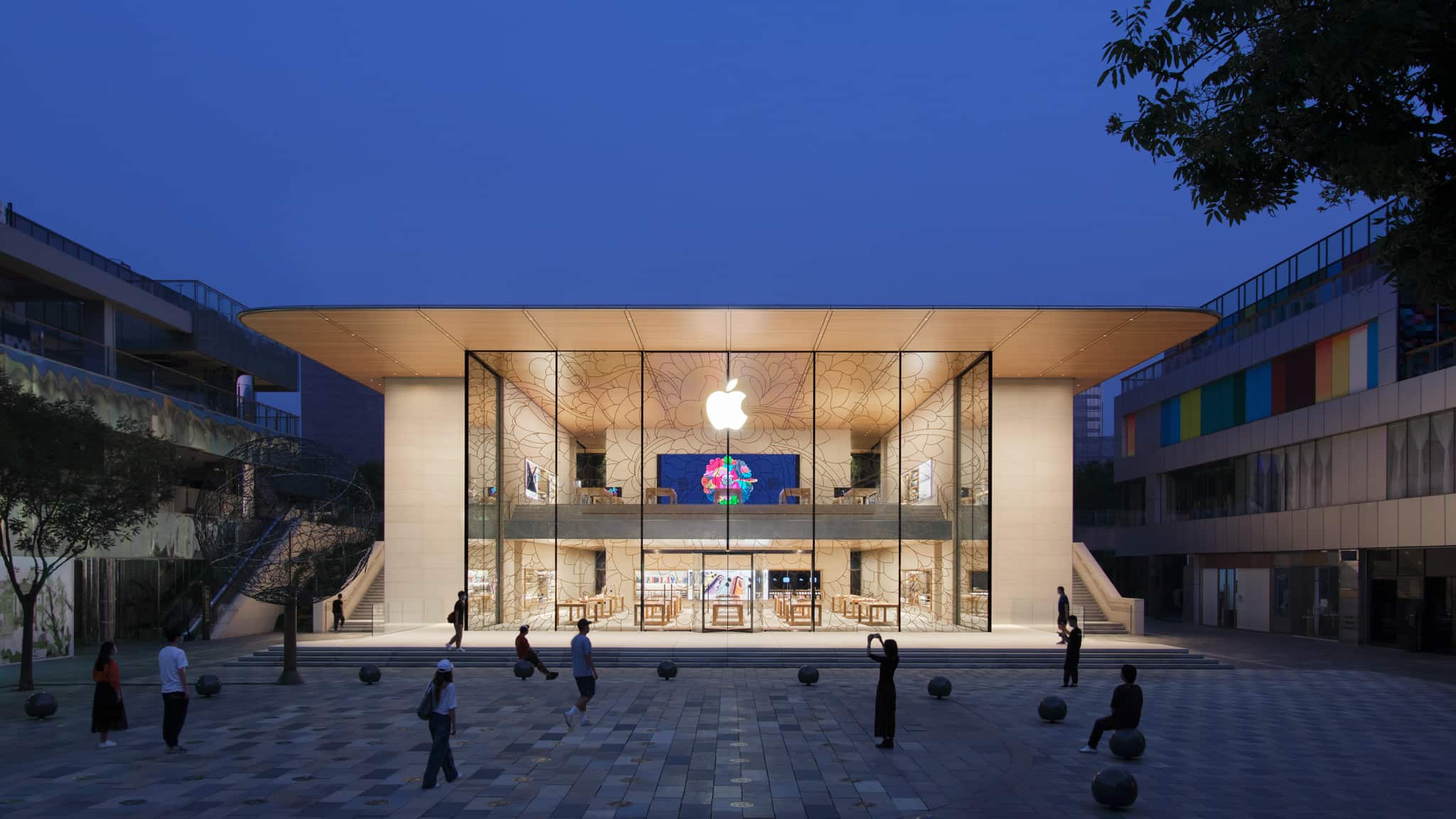 Exterior view of Apple Sanlitun featuring a pavilion design, which envelopes a 2-storey tall space with a 10 meter tall glass store front facing a plaza and a pair of stairs on either side. The store’s timber ceiling is melded into a complex double curved stainless steel roof paved with photovoltaic panels that generates electricity for the store’s usage.