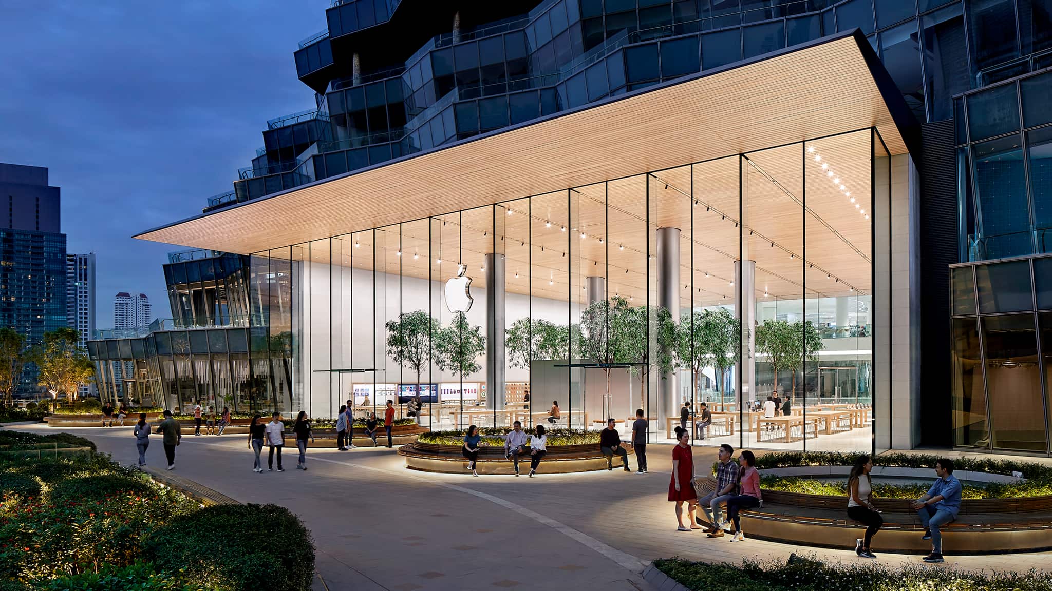 Angled exterior view of Apple Iconsiam featuring a 10 meter glass facade with a sloping timber slat ceiling extends outward as a 6 meter cantilevered canopy along the exterior. The store is elevated above the river bank, providing scenic views and customer access to a lush roof terrace.